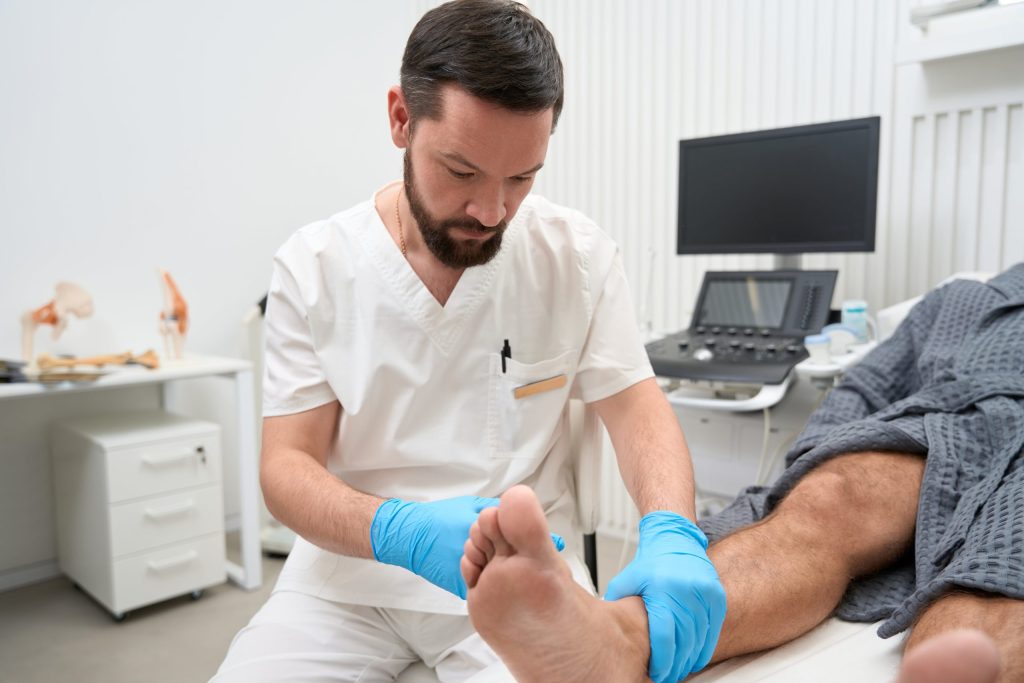 A Focused Male Doctor Examines A Patient's Foot In A Medical Office, Wearing Blue Gloves For A Thorough Foot Assessment.