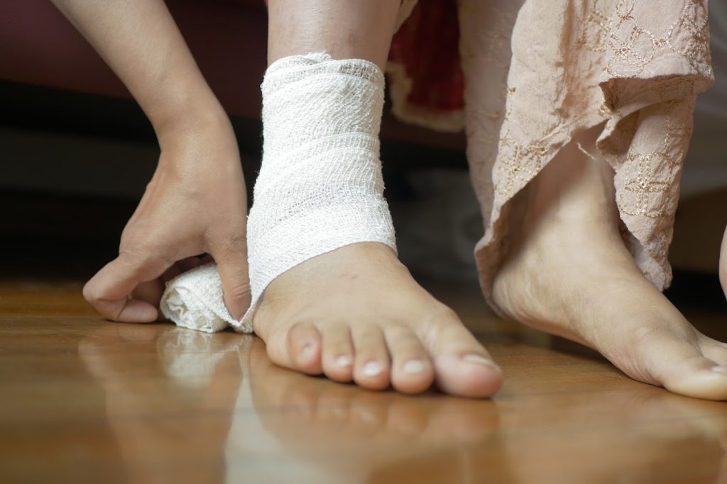 Woman With Bandaged Foot On Wooden Floor, Adjusting Wrap For Injury.